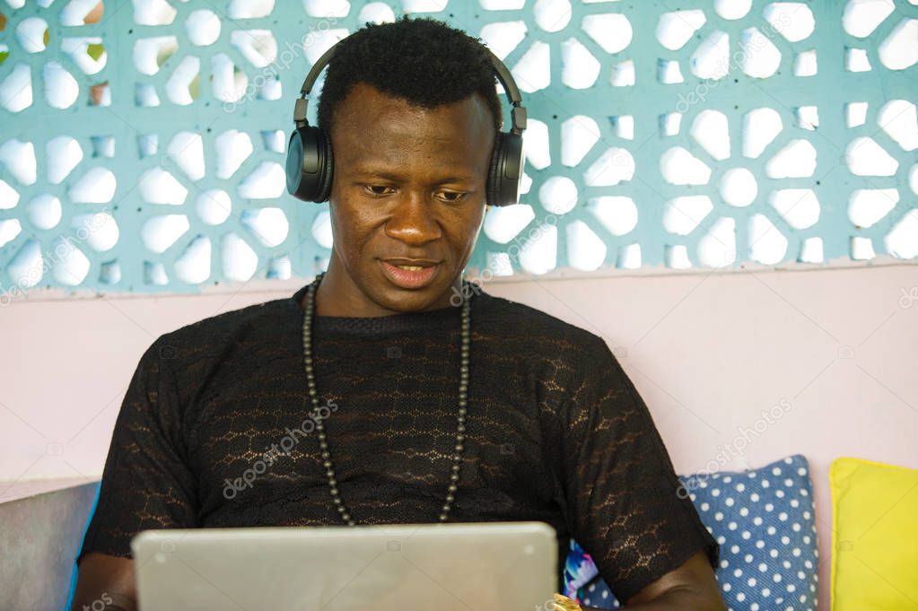 young handsome and happy hipster black African American business man working with laptop computer and headphones smiling satisfied and cool in digital nomad