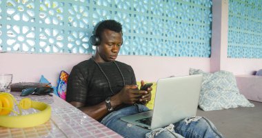 Dijital göçebe iş cep telefonu ve dizüstü bilgisayar ile rahat kulaklık ağ ile genç yakışıklı ve serin siyah Afrikalı Amerikan hipster adam