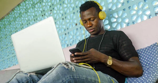 cool and attractive black afro American hipster man with headphones networking relaxed with mobile phone and laptop computer in digital nomad job and internet work