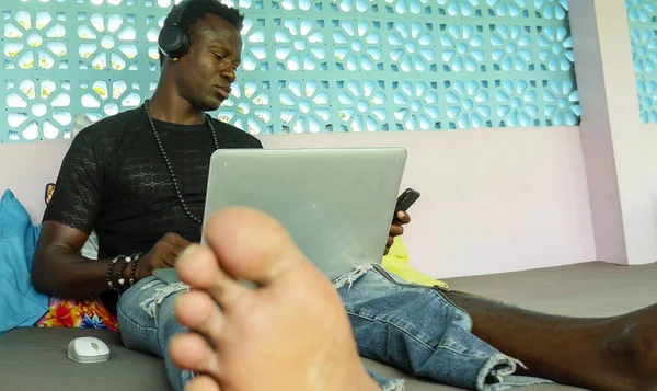 Νέος όμορφος και δροσερό μαύρο Αφρικανός Αμερικανός άντρας με ακουστικά δικτύωσης χαλαρή με το κινητό τηλέφωνο και φορητό υπολογιστή στην εργασία ψηφιακών νομάδων — Φωτογραφία Αρχείου