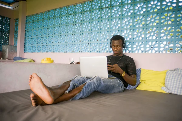 Молодой красивый и классный черный афроамериканец хипстер с наушниками, расслабленный с мобильным телефоном и ноутбуком в цифровой кочевой работе — стоковое фото