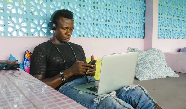 Joven guapo y fresco negro afroamericano hipster hombre con auriculares de red relajado con el teléfono móvil y ordenador portátil en el trabajo nómada digital — Foto de Stock