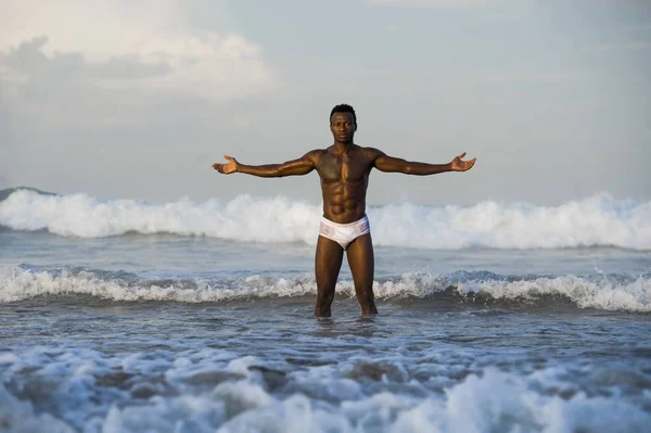 artistic portrait of young attractive and fit black African American man with sexy muscular body and six pack abs opening arms free on beach with big sea waves