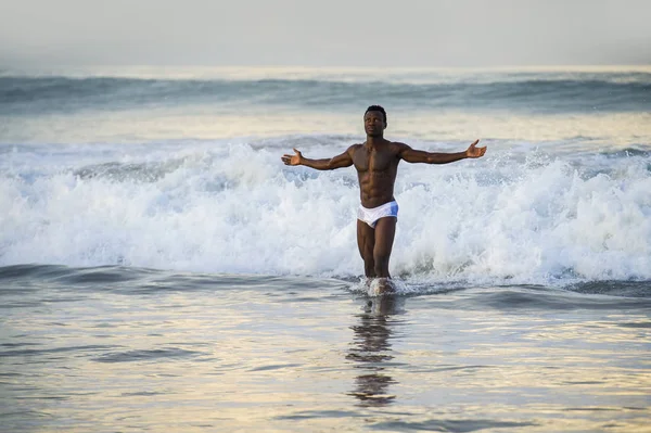 artistic portrait of young attractive and fit black afro American man with sexy muscular body and six pack abs opening arms free on beach with big sea waves