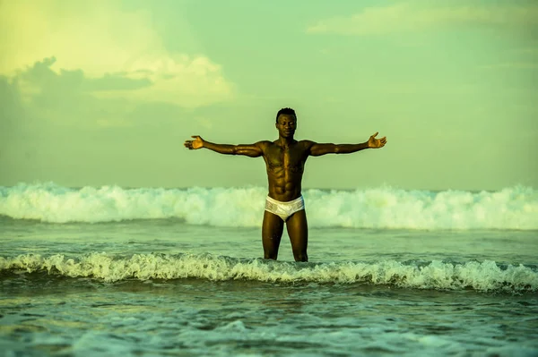 artistic portrait of young attractive and fit black afro American man with sexy muscular body and six pack abs opening arms free on beach with big sea waves