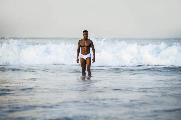 attractive and fit black African American man with sexy muscular body and sixpack abs posing cool on beach with big sea waves and foam around in beauty and health concept