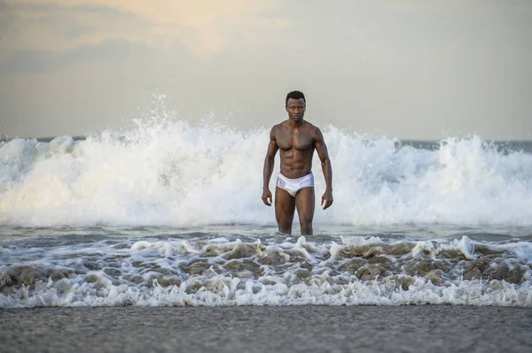 attractive and fit black African American man with sexy muscular body and sixpack abs posing cool on beach with big sea waves and foam around in beauty and health concept
