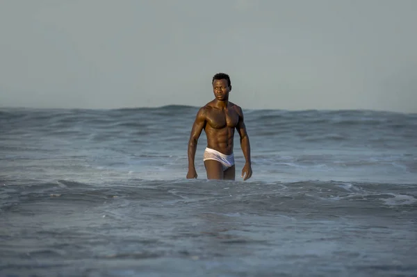 attractive and fit black African American man with sexy muscular body and sixpack abs posing cool on beach with big sea waves and foam around in beauty and health concept