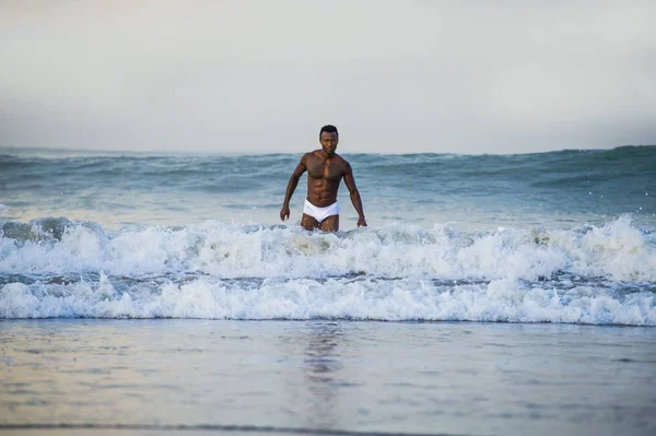 attractive and fit black afro American man with sexy muscular body and sixpack abs posing cool on beach with big sea waves and foam around in beauty and health concept