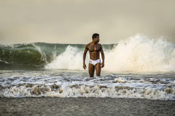 attractive and fit black afro American man with sexy muscular body and sixpack abs posing cool on beach with big sea waves and foam around in beauty and health concept