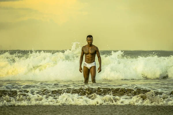 attractive and fit black afro American man with sexy muscular body and sixpack abs posing cool on beach with big sea waves and foam around in beauty and health concept