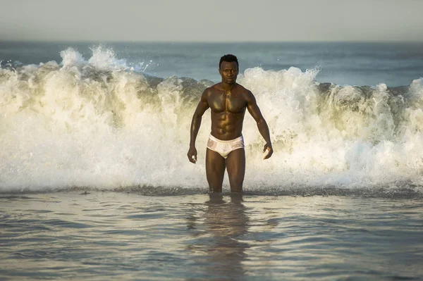 attractive and fit black afro American man with sexy muscular body and sixpack abs posing cool on beach with big sea waves and foam around in beauty and health concept