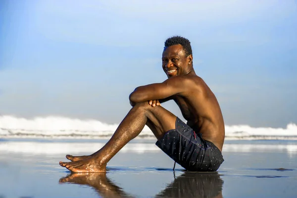 Joven atractivo y feliz negro afro americano hombre con ajuste muscular cuerpo sentado en la playa arena disfrutando hermosa vista sonriente alegre en vacaciones de verano — Foto de Stock