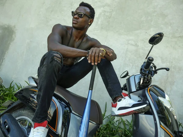 urban lifestyle portrait of fit body and dangerous looking black afro American man with naked torso and sunglasses sitting on scooter holding baseball bat