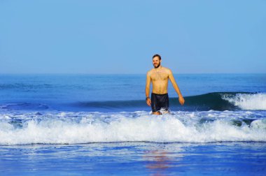 Genç, çekici, sakallı ve yüzme şortlu adam tropikal cennet kumsalında yalnız başına neşeli ve neşeli bir şekilde yaz tatilinin tadını çıkarıyor.