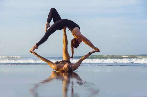 Young attractive and beautiful acrobat couple practicing acroyoga exercise concentrated keeping balance practiing yoga on the beach in relaxation and meditation concept — Stock Photo, Image
