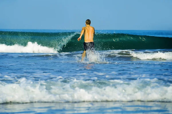 Pohled na mladého atraktivního a šťastného muže v plaveckých paškách na pouštní pláži tropický ráj, hravý běh k mořské vodě hrající velkými vlnami na dovolenou — Stock fotografie