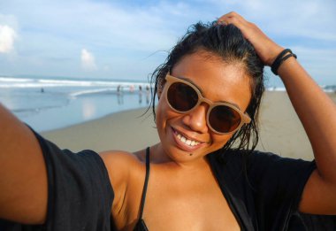 Genç, mutlu ve çekici Asyalı egzotik kadın sahilde cep telefonuyla fotoğraf çekiyor neşeli gülümseyerek güzel tropikal adada tatilin tadını çıkarıyor.