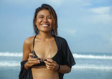 Genç egzotik ve çekici Asyalı kız elinde güneş gözlüğü ile tropik sahilde tatilin tadını çıkarıyor mutlu ve kaygısız bir şekilde denizde eğleniyor.