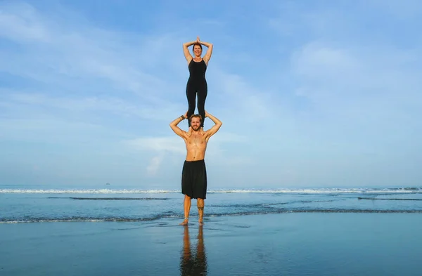 Молодая привлекательная и красивая пара акробатов практикующих акройогу упражнения сосредоточены поддержания баланса практикующих йогу на пляже в концепции релаксации и медитации — стоковое фото