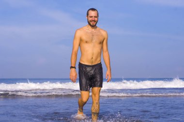 Genç, çekici, sakallı ve yüzme şortlu adam tropikal cennet kumsalında yalnız başına neşeli ve neşeli bir şekilde yaz tatilinin tadını çıkarıyor.