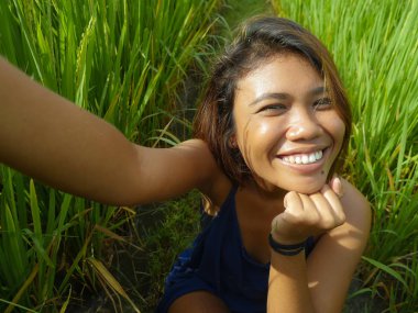 Mutlu ve egzotik adalı Endonezyalı genç bir kız cep telefonuyla kendi portresini çekiyor neşeli ve heyecanlı bir şekilde pirinç tarlası arka planında poz veriyor.