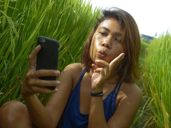Young happy and exotic islander Asian girl from Indonesia taking selfie self portrait photo with mobile phone smiling cheerful and excited posing rice field nature background — Stock Photo, Image