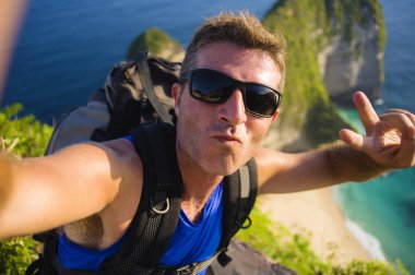 Genç, mutlu ve çekici sırt çantalı adam cep telefonuyla güzel sahil kayalıklarında gülümseyen neşeli deniz manzarasının önünde selfie portresi çekiyor.