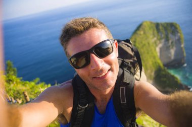 Genç, mutlu ve çekici sırt çantalı adam cep telefonuyla güzel sahil kayalıklarında gülümseyen neşeli deniz manzarasının önünde selfie portresi çekiyor.