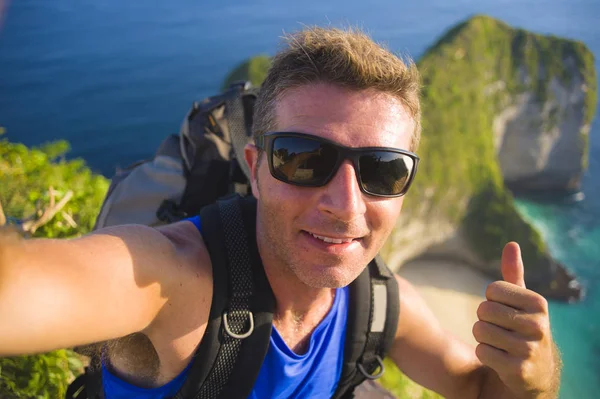 Ung glad och attraktiv backpacker man tar selfie porträtt foto med mobiltelefon framför fantastiska havet landskap ler glad vandring på vackra strand klippa — Stockfoto