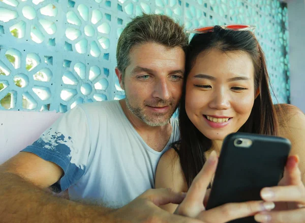Смешанная этническая пара влюблена улыбается весело с красивым кавказским мужчиной и красивой азиатской китаянкой, используя мобильный телефон вместе в кофейне — стоковое фото