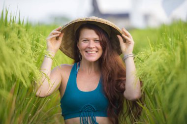 Geleneksel Asyalı çiftçi şapkalı, mutlu ve güzel kızıl saçlı kadın Asya 'da yeşil pirinç tarlasında izole edilmiş neşeli bir şekilde gülümsüyor. Turist gezisi ve yaz tatili.