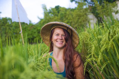 Geleneksel Asyalı çiftçi şapkalı, mutlu ve güzel kızıl saçlı kadın Asya 'da yeşil pirinç tarlasında izole edilmiş neşeli bir şekilde gülümsüyor. Turist gezisi ve yaz tatili.