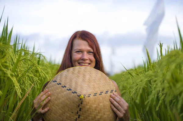 Young happy and beautiful red hair woman playing with traditional Asian farmer hat smiling cheerful isolated on green rice field in Asia tourist trip and Summer holidays — Stock Photo, Image