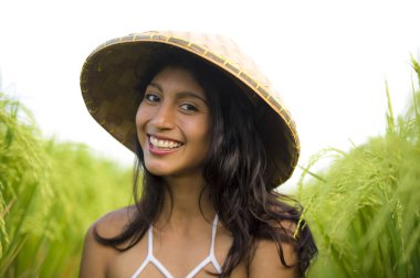 young happy and beautiful latin woman playing with traditional Asian farmer hat smiling having fun posing sexy isolated on green rice field in Asia tourist trip clipart