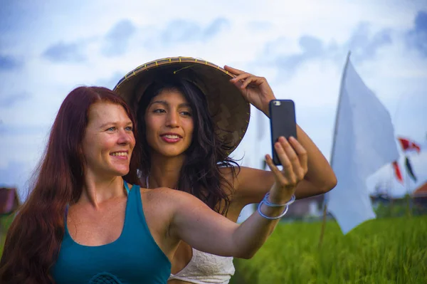 Geleneksel Asyalı çiftçi şapkalı genç ve güzel İspanyol kız ve Kafkasyalı kadın pirinç tarlasında cep telefonuyla kız arkadaş selfie 'si çekiyorlar. — Stok fotoğraf