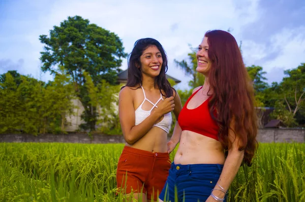 Genç ve güzel Latin kız ve onun çekici kızıl saçlı kız arkadaşı yaz tatilinin tadını çıkarıyorlar pirinç tarlasında yürüyorlar mutlu bir şekilde gülümsüyorlar etnik çeşitlilikte rahatlamış olarak. — Stok fotoğraf