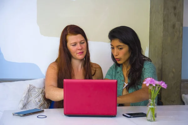 Jóvenes felices y hermosas mujeres caucásicas e hispanas que trabajan en la cafetería de la oficina con computadora portátil como socios de negocios digitales y amigos étnicos diversidad — Foto de Stock