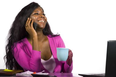 Genç, mutlu ve çekici Afro-Amerikalı iş kadını kahve içiyor. Ofis masasında rahatça çalışıyor ve cep telefonuyla konuşuyor.