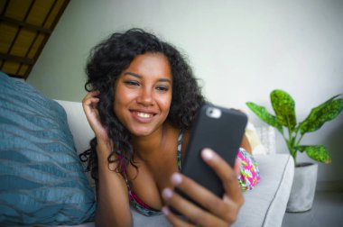 Genç, güzel ve mutlu, karışık etnik kökenli, beyaz ve siyah afro Amerikalı bir kadın cep telefonu uygulaması kullanarak evinde uzanıyor ve neşeli bir şekilde gülüyor.