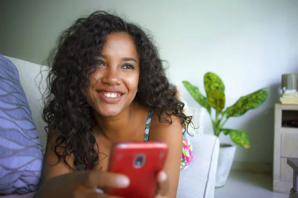 Νεαρή όμορφη και ευτυχισμένη μικτή εθνικότητα Καυκάσια και μαύρη Αφρικανή Αμερικανίδα ξαπλωμένη στον καναπέ του σπιτιού χρησιμοποιώντας εφαρμογή κινητού τηλεφώνου που χρονολογείται online χαμογελώντας χαρούμενα — Φωτογραφία Αρχείου