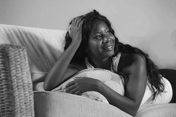 Estilo de vida casa retrato de joven hermosa y feliz negro afro mujer americana acostada en el sofá de la sala de estar sonriendo alegre como si soñara despierto en blanco y negro — Foto de Stock