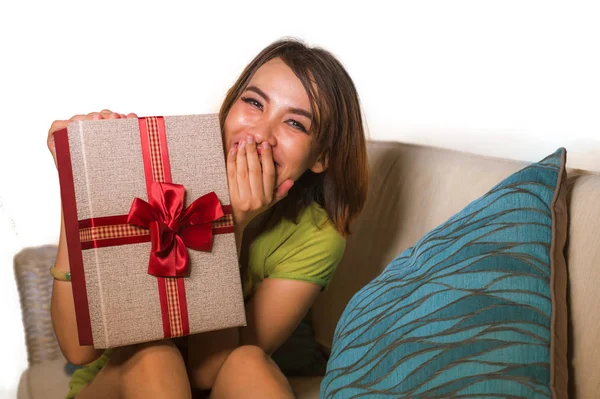 Νεαρή όμορφη και ευτυχισμένη ασιατική ινδονησιακή γυναίκα που κατέχουν γενέθλια ή δώρο Χριστουγέννων δείχνει το κουτί δώρου χαρούμενα και ενθουσιασμένος κάθεται στο καναπέ του σαλονιού — Φωτογραφία Αρχείου