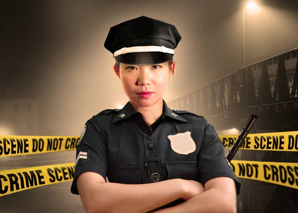 young Asian American police officer standing serious in custody of crime scene for preserving evidence at do not cross police line yellow tape in murder investigation