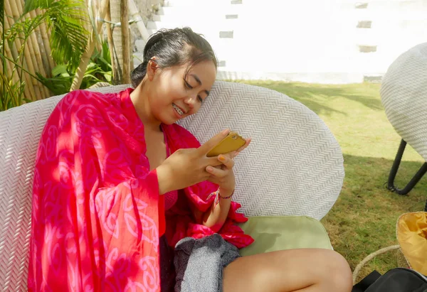 Joven feliz y hermosa asiática adolescente indonesia en bikini sonriendo en la cama de la piscina con sarong usando aplicación de medios sociales en internet teléfono móvil relajado en el complejo tropical —  Fotos de Stock