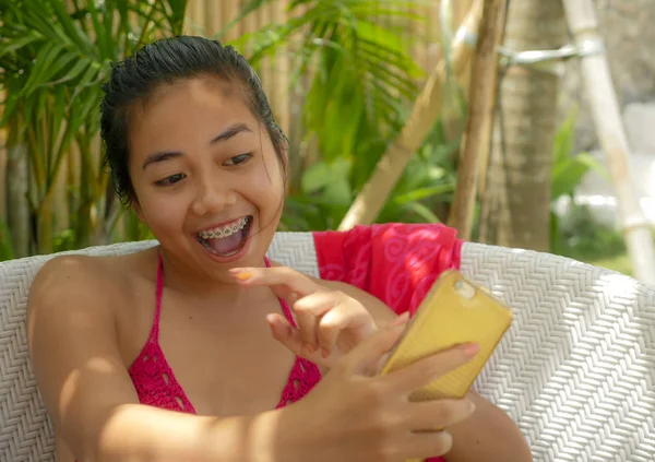 Young happy and beautiful Asian Indonesian teenager girl in bikini sitting on pool bed with sarong taking selfie picture with mobile phone relaxed at tropical resort — Stock Photo, Image