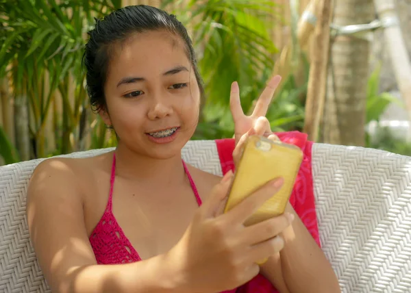 Joven feliz y hermosa chica adolescente indonesia asiática en bikini sentado en la cama de la piscina con sarong tomar foto selfie con teléfono móvil relajado en el complejo tropical —  Fotos de Stock