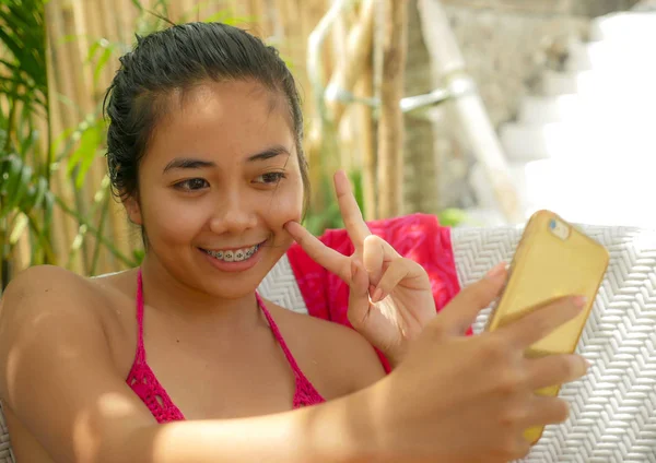 Молода щаслива і красива індонезійська дівчина підліткового віку в бікіні сидить на ліжку в басейні, а Саронг робить фотографії з мобільним телефоном, розслабленим на тропічному курорті. — стокове фото