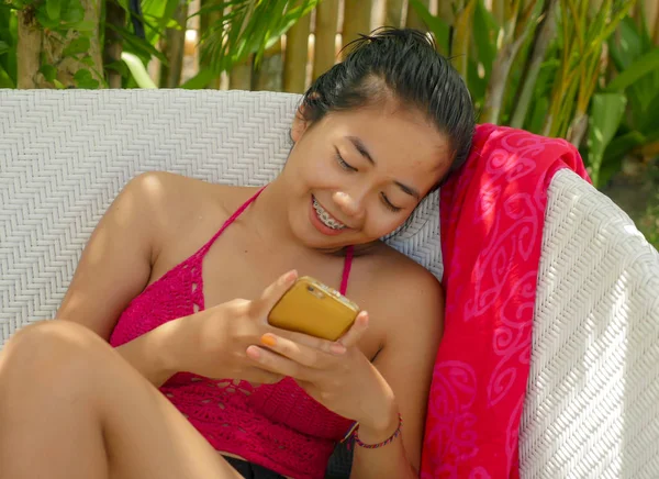 Young happy and beautiful Asian Indonesian teenager girl in bikini smiling on pool bed with sarong using social media app on internet mobile phone relaxed at tropical resort — 스톡 사진