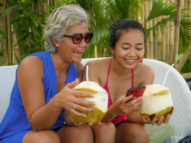 mutlu ve güzel Asya Endonezyalı genç kız annesi ile birlikte tropikal tatil bahçesinde hindistan cevizi içme Yaz tatili zevk cep telefonu kullanarak